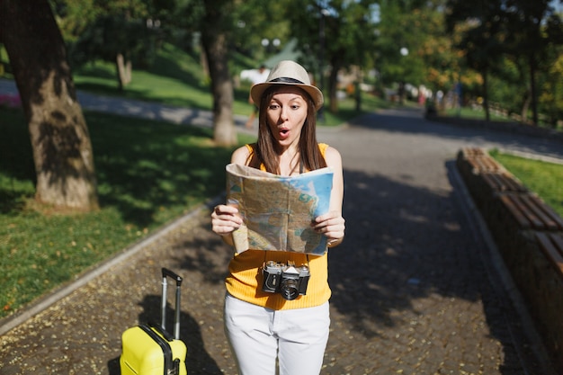 Schockierte Reisende Touristenfrau in gelber Sommerkleidung, Hut mit Koffer, der auf Stadtplan in der Stadt im Freien schaut. Mädchen, das ins Ausland reist, um am Wochenende zu reisen. Tourismus Reise Lebensstil.