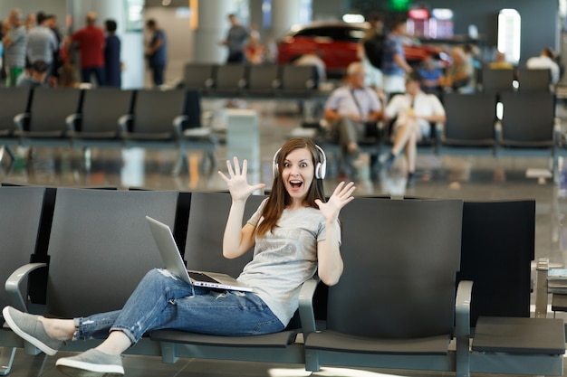 Schockierte reisende Touristenfrau mit Kopfhörern, die Musik hört, am Laptop arbeitet, die Hände ausbreitet, in der Lobbyhalle am Flughafen wartet?