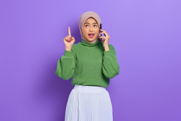 Schockierte schöne asiatische Frau in grünem Pullover und Hijab, die auf dem Handy spricht und mit dem Finger nach oben zeigt, wodurch geniale Lösungen isoliert über violettem Hintergrund entstehen