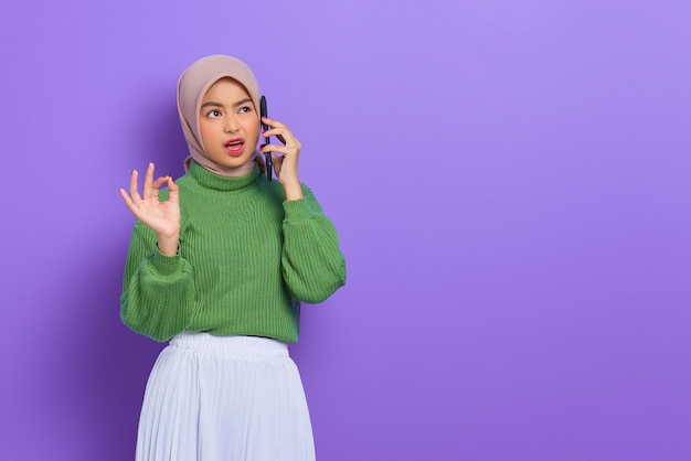 Schockierte schöne asiatische Frau in grünem Pullover und Hijab, die auf dem Handy telefoniert und ein Ok-Zeichen gestikuliert, isoliert vor violettem Hintergrund