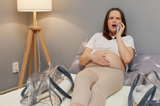 Schockierte schwangere Frau, die schmerzhafte Kontraktionen und starke Schmerzen fühlt, mit dem Smartphone spricht, den Arzt ruft einen Krankenwagen an, berührt ihren Bauch und macht Grimassen vor Schmerzen