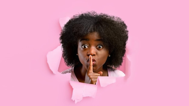 Schockierte tausendjährige schwarze Frau schaut in Loch in rosa Papier und zeigt Finger in der Nähe von Lippen Zeichen der Stille