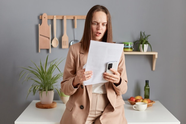 Schockierte, überraschte, enttäuschte Frau mit braunen Haaren, die eine beige Jacke trägt, die in der Küche zu Hause steht, mit einem Smartphone in den Händen, das Papiere mit Erstaunen liest