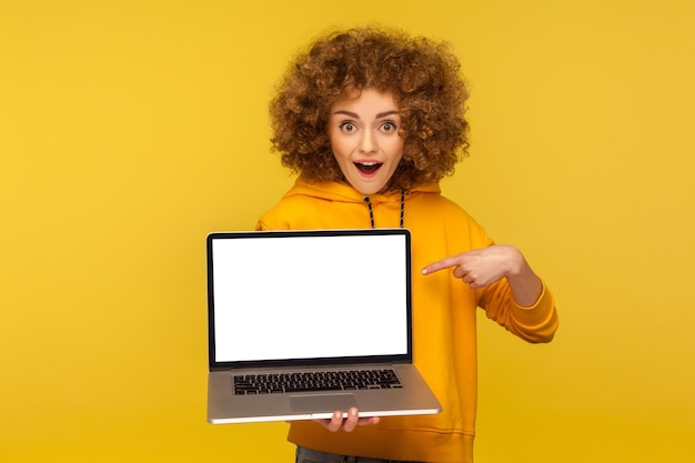 Schockierte, überraschte Frau mit flauschigem, lockigem Haar im Kapuzenpullover im urbanen Stil, die auf einen leeren Laptop-Bildschirm zeigt und erstaunt aussieht, als sie ein leeres Mock-up-Display für eine isolierte Studioaufnahme einer Webanzeige zeigt