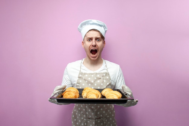 Schockierter Bäcker in Uniform hält frisch gebackene Croissants in der Hand und schreit mit offenem Mund