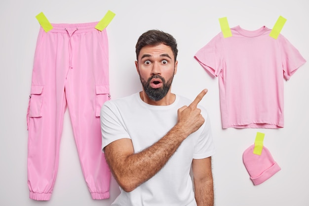 Schockierter bärtiger erwachsener mann zeigt auf verputzte kleidung, hat einen erstaunten ausdruck und zeigt gegenstände zum tragen von posen vor weißem hintergrund mit verputzten hosen, t-shirt und hut. omg schau dir das an