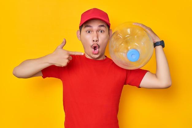 Schockierter junger asiatischer Mann in roter Kappe, T-Shirt, einheitliche Mitarbeiterarbeit als Händlerkurier, der mit dem Finger auf eine Wasserflasche zeigt, die auf gelbem Hintergrund isoliert ist Professionelles Lieferservice-Konzept