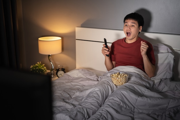 Schockierter junger Mann, der nachts einen Fernsehfilm auf einem Bett sieht