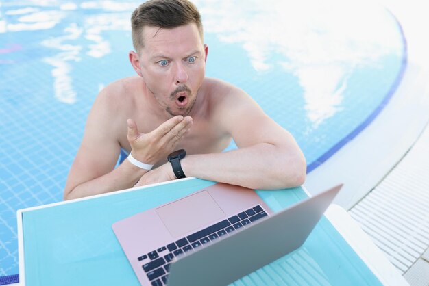 Schockierter junger Mann im Pool schaut in einen Laptop