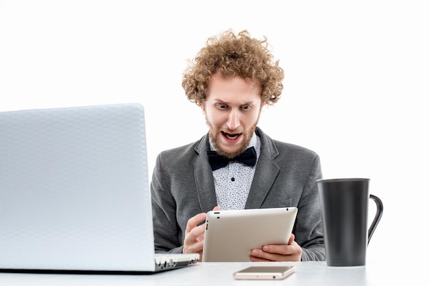 Schockierter lockiger Geschäftsmann, der gegen einen Laptop schreit, während er ein digitales Tablet benutzt