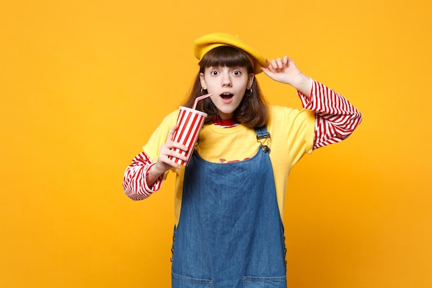 Schockierter Mädchen-Teenager in französischer Baskenmütze, Denim-Sommerkleid halten Plastikbecher mit Cola oder Limonade, die den Mund offen halten, isoliert auf gelbem Hintergrund. Menschen aufrichtige Emotionen, Lifestyle-Konzept. Mock-up-Kopienbereich.