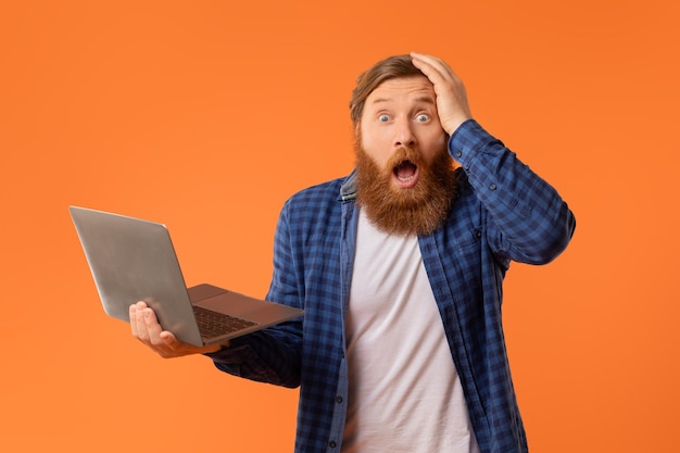 Schockierter rothaariger bärtiger Mann, der mit einem Laptop posiert, berührt das Studio.