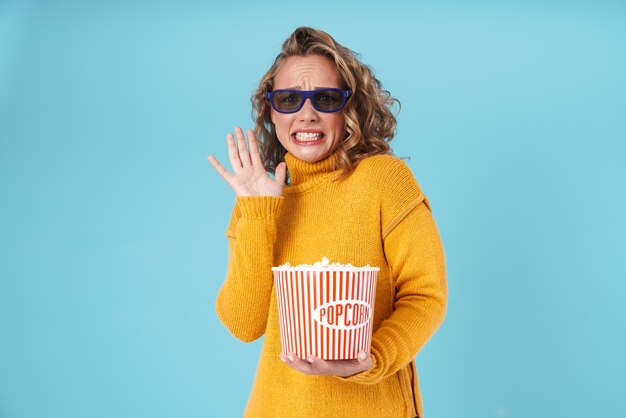 Schockiertes hübsches junges Mädchen, das einen Pullover trägt, der isoliert auf Blau steht und Popcorn isst, während er einen Film sieht