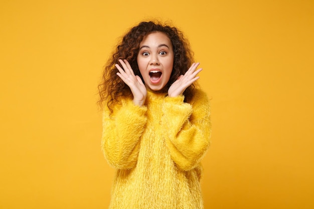 Schockiertes junges afrikanisch-amerikanisches Mädchen im Pelzpullover posiert isoliert auf gelb-orangefarbenem Wandhintergrund, Studioporträt. People-Lifestyle-Konzept. Mock-up-Kopienbereich. Mund offen halten, Hände ausbreiten.