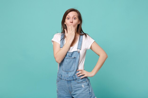 Schockiertes junges brünettes Frauenmädchen in der zufälligen Denimkleidung, die lokalisiert auf blauem Türkiswandhintergrundstudioporträt aufwirft. Menschen Lifestyle-Konzept. Kopieren Sie Platz. Mund mit der Hand bedecken.