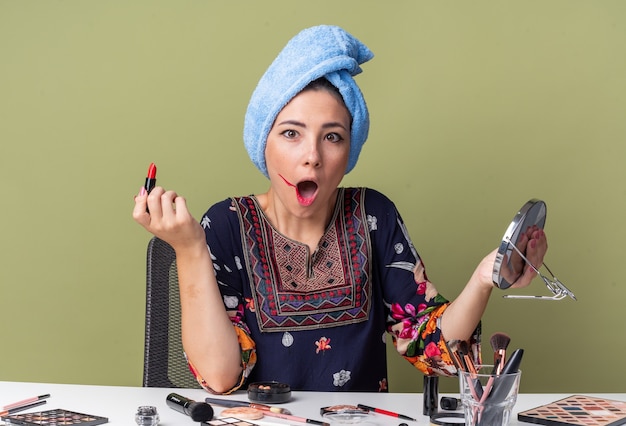 Foto schockiertes junges brünettes mädchen mit eingewickelten haaren im handtuch, das am tisch mit make-up-tools sitzt, die spiegel und lippenstift halten, isoliert auf olivgrüner wand mit kopierraum