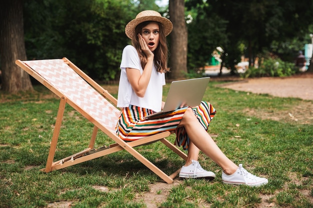 Schockiertes junges Mädchen, das mit Laptop-Computer am Park draußen sitzt