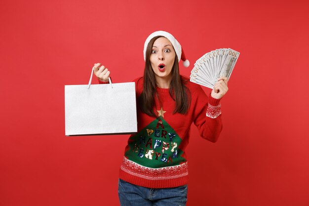 Schockiertes junges Santa-Mädchen hält viele Dollar-Banknoten Bargeld, verpackt Taschen mit Einkäufen nach dem Einkaufen einzeln auf rotem Hintergrund. Frohes neues Jahr 2019 Feier Urlaub Party Konzept.