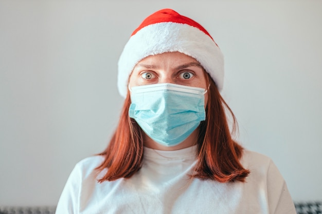 Schockiertes kaukasisches Mädchen mit Weihnachtsmütze und medizinischer Schutzmaske schaut in den Camet