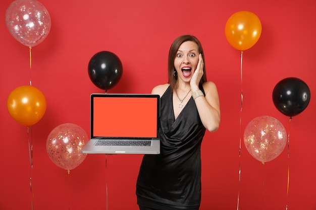 Schockiertes Mädchen im schwarzen Kleid, das die Hand auf dem Gesicht hält, das Laptop-PC-Computer mit leerem schwarzem leerem Bildschirm auf hellen roten Hintergrundluftballons hält. Frohes neues Jahr, Geburtstagsmodell Urlaub Partykonzept.