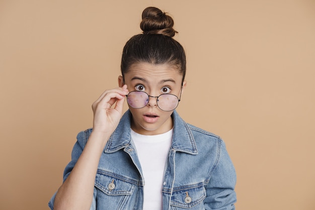 Schockiertes Mädchen mit weggenommenen Haaren schaut überrascht durch ihre Brille. Kleines Mädchen auf braunem Hintergrund