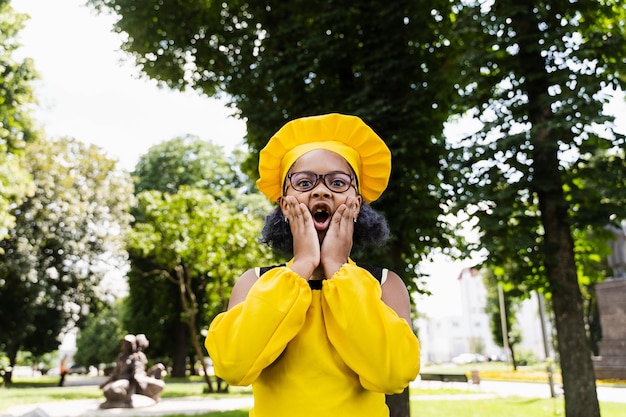 Schockiertes schwarzafrikanisches Kinderkochmädchen in Kochmütze und gelber Schürzenuniform halten Wangen und überraschen Kreative Werbung für Café oder Restaurant