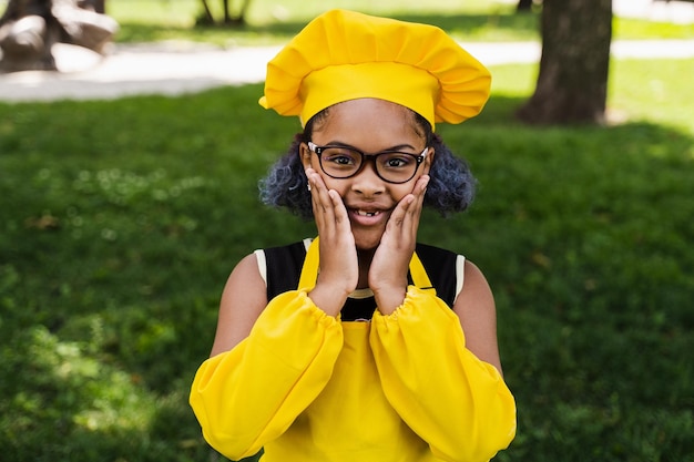 Schockiertes schwarzafrikanisches Kinderkochmädchen in Kochmütze und gelber Schürzenuniform halten Wangen und überraschen Kreative Werbung für Café oder Restaurant