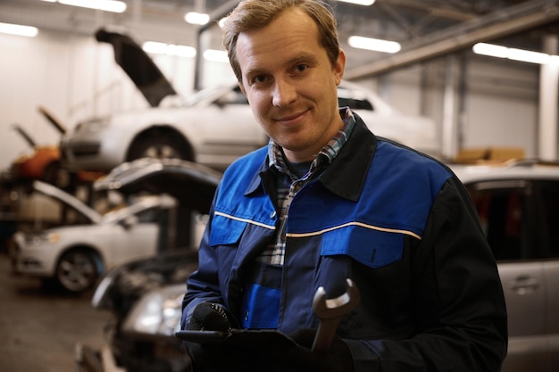 Schön lächelnder kaukasischer Automechaniker, Autoingenieur, Techniker in Uniform, der einen Schraubenschlüssel hält und in der Werkstatt gegen angehobene Autos mit offener Motorhaube steht