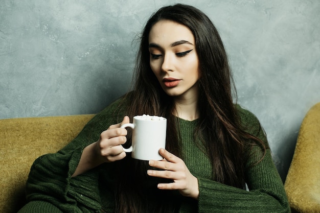 Schön und süß Schöne junge Frau, die eine Kaffeetasse hält und zu Hause auf dem Stuhl sitzt Lifestyle-Konzept