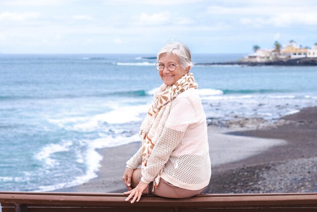 Schöne ältere Frau, die eine Brille trägt, die auf einer Bank im Freien auf dem Meer sitzt und die Kamera betrachtet, die Freiheit und Urlaub genießt Aktive kaukasische ältere Frau, die sich an einem sonnigen Tag in der Nähe des Strandes entspannt