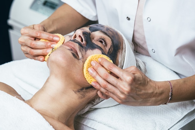 Schöne ältere Frau, die eine Tongesichtsmaske mit verjüngenden Effekten im Spa-Schönheitssalon erhält.