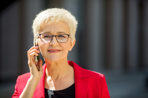 schöne ältere Frau im Alter mit einem kurzen Haarschnitt und Brille in einer roten Jacke mit einem Telefon