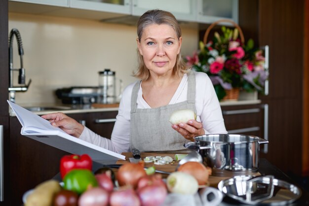 Foto schöne ältere frau in der schürze liest ein buch der rezepte, während in der küche kochend