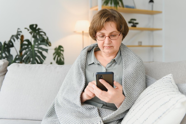 Schöne ältere Frau in Freizeitkleidung mit ihrem Smartphone beim Sitzen auf dem Sofa zu Hause