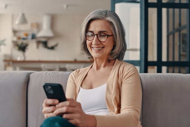 Schöne ältere Frau in Freizeitkleidung mit ihrem Smartphone beim Sitzen auf dem Sofa zu Hause