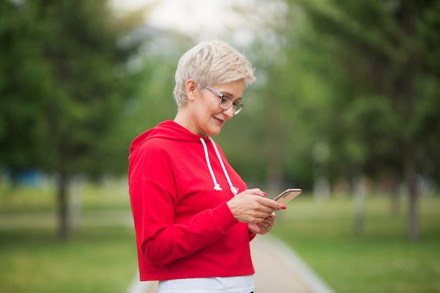 schöne ältere Frau in Sportbekleidung mit einem Telefon in der Hand im Park