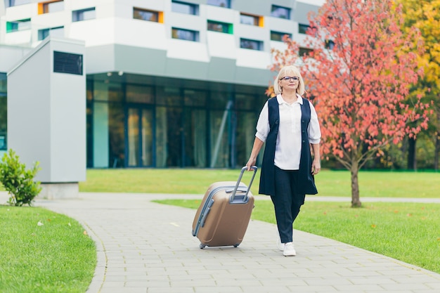 Schöne ältere Frau reist mit einem großen Reisekoffer, auf dem Hintergrund eines schönen modernen Gebäudes