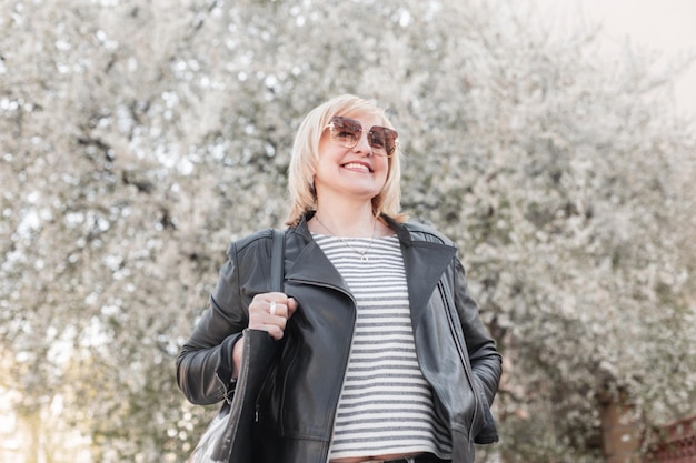 Schöne ältere glückliche modische Frau mit Sonnenbrille in modischer Kleidung und einer Lederjacke und stilvoller Handtasche geht spazieren und genießt in einem Park mit blühenden Bäumen