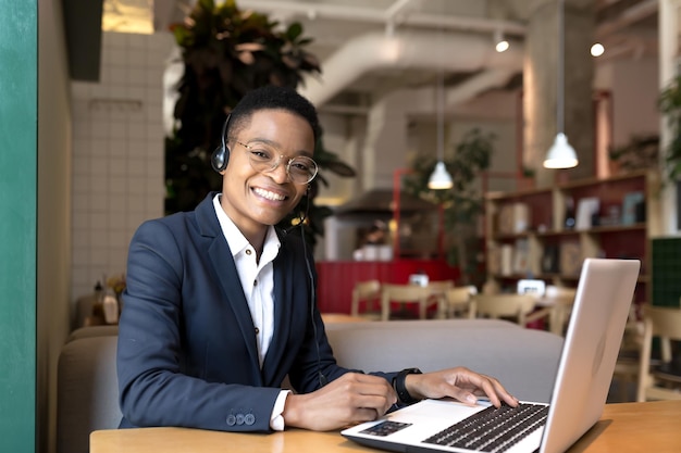 Schöne afrikanisch-amerikanische Frau, die in die Kamera schaut und lächelt, arbeitet in der Videokommunikation eines Coworking-Cafés mit einem Headset, einer freiberuflichen Geschäftsfrau