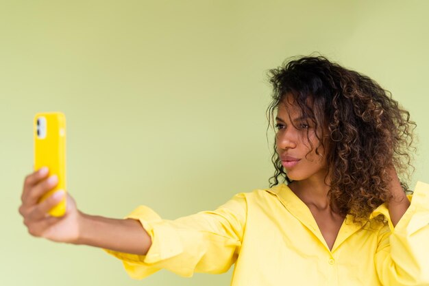 Schöne afrikanisch-amerikanische Frau im Freizeithemd auf grünem Hintergrund, die gerne ein Selfie auf dem Smartphone macht