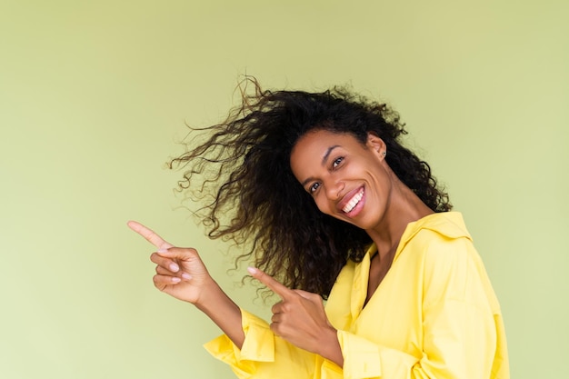 Schöne afrikanisch-amerikanische Frau im Freizeithemd auf grünem Hintergrund, glücklicher positiver aufgeregter Zeigefinger nach links