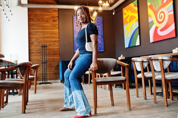 Schöne afrikanisch-amerikanische Frau mit Dreadlocks in blauen, stylischen Jeans im Café Schöne coole modische schwarze junge Mädchen drinnen