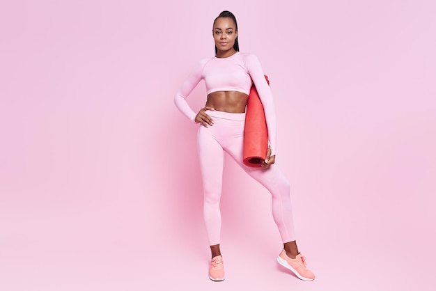 Schöne afrikanische Frau in Sportkleidung trägt eine Trainingsmatte auf rosa Hintergrund