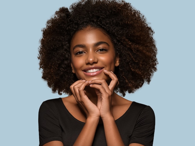 Schöne afrikanische Frau schwarzes T-Shirt Porträt Afro-Haarschnitt, der ihr Gesicht berührt Farbhintergrund blau