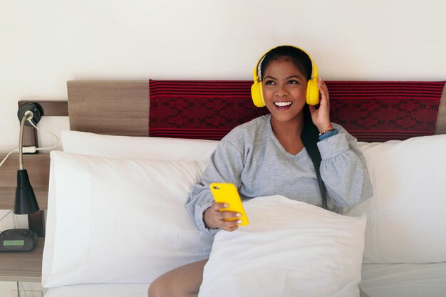Schöne Afroamerikanerin, die auf ihrem Bett in ihrem Pyjama sitzt und Musik mit ihrem Handy und Kopfhörern hört.