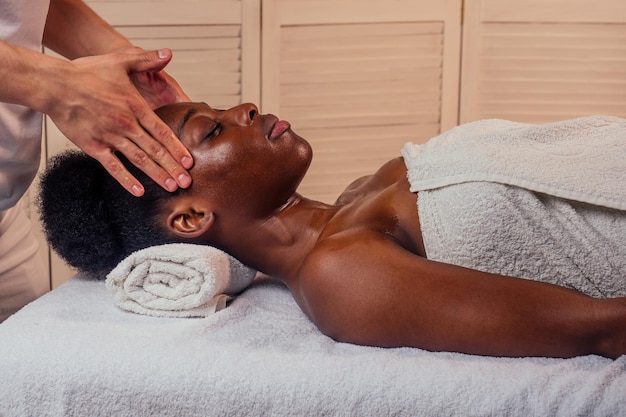 Schöne afroamerikanische Frau, die Ayurveda-Gesichtsmassage durch die weißhäutigen Hände des Mannes im Spa-Salon bekommt