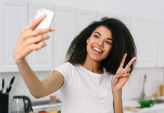 Schöne afroamerikanische Frau, die Handy verwendet, Selfie nimmt, Siegeszeichen zeigend. Blogger-Streaming-Video online