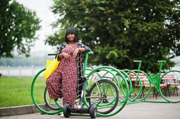 Schöne afroamerikanische Frau, die in der Nähe von Segway oder Hoverboard und Fahrrad steht. Schwarzes Mädchen mit gelben Stoff-Eco-Taschen, die Symbol recyceln.