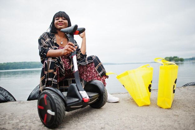 Schöne afroamerikanische Frau sitzt in der Nähe von Segway oder Hoverboard. Schwarzes Mädchen mit gelben Stoff-Eco-Taschen, die Symbol recyceln.