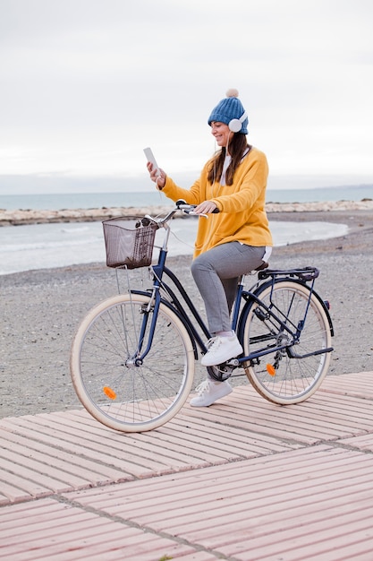 Schöne aktive reife Frau mit Fahrrad auf der Straße durch das Meer. Glücklich lächelnde Frau mittleren Alters mit Kopfhörern, die Musik hören, kopieren Raum